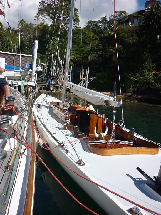 A white sailboat with wooden trim, reminiscent of Classic Sailing Yachts, is moored beside another boat. The sail is furled and the boat rests on calm water, surrounded by trees and waterfront structures.
