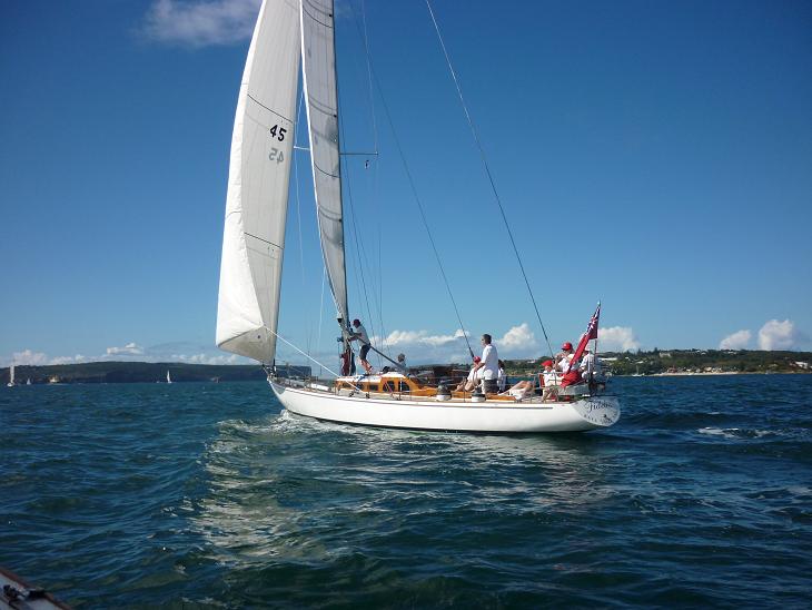 A white Square Metre Yacht with several people on board sails on blue water under a clear sky, with a coastline and green hills visible in the background.