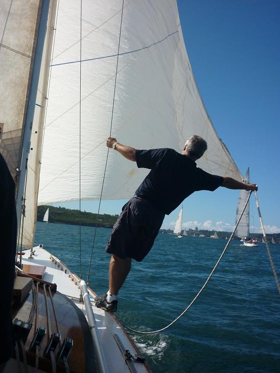 A man balances on the edge of a sailboat, holding a rope whilst adjusting the sail of a classic sailing yacht on a sunny day. Other Skerry Cruisers and the green shoreline appear in the background under a blue sky.