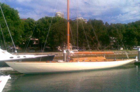 A classic wooden Schärenkreuzer with a tall mast is moored at a marina, floating on calm water, with trees and buildings visible in the background.