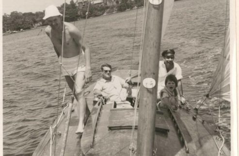 Four people relax on a classic sailing yacht: one man stands shirtless near the bow, while three others—including a child—sit behind him. Trees and buildings line the shore. The caption reads MARCH 1962.
