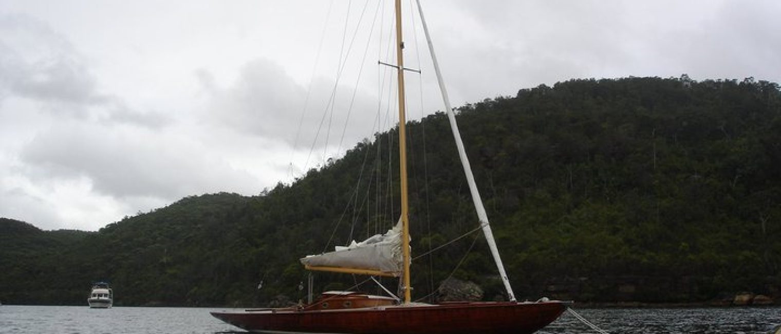 A wooden Skerry Cruiser with its sail down is anchored on calm water, forested hills rising in the background beneath an overcast sky. Another boat is visible in the distance.