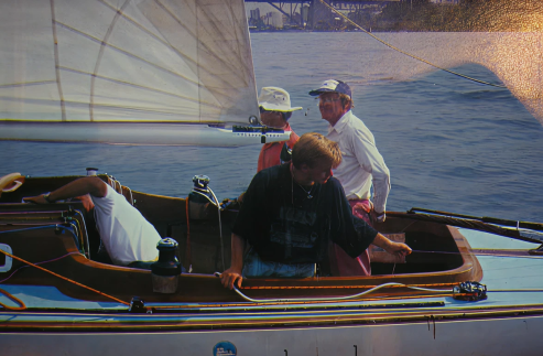 Four people on a Classic Sailing Yacht are preparing for sailing. One person is bent over inside the boat, whilst the others stand or sit, gazing in different directions. The water and a distant bridge form the scenic backdrop.