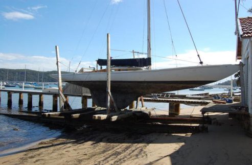 A white Classic Sailing Yacht is positioned on wooden supports at a boatyard, partially out of the water, with a quay and several boats visible in the background under a clear sky.