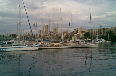 Several Square Metre Yachts and classic sailing yachts are moored in a marina, with tall buildings and flats in the background under a cloudy sky. The serene scene is beautifully reflected on the calm water below.