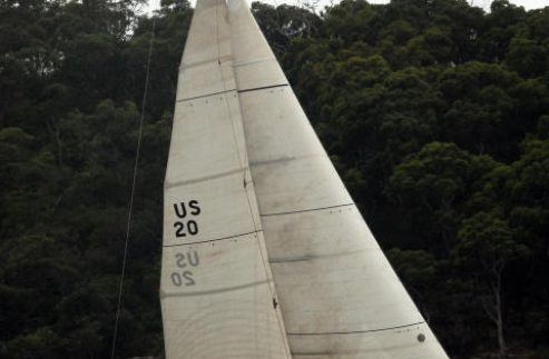 A classic Square Metre Yacht with white sails marked US 20 glides on choppy water near a forested shoreline under an overcast sky. Several people are visible on board, adjusting the sails.