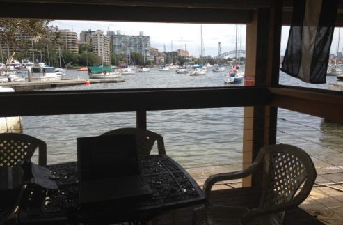 A shaded patio with a table, laptop, and chairs overlooks a marina featuring elegant Skerry Cruisers moored on the water. City buildings and a bridge rise in the background beneath a partly cloudy sky.