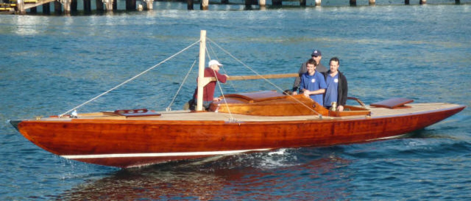 A polished wooden motorboat with three people aboard glides on blue water near a white pier and green, tree-lined shore, passing elegant Square Metre Yachts moored nearby.