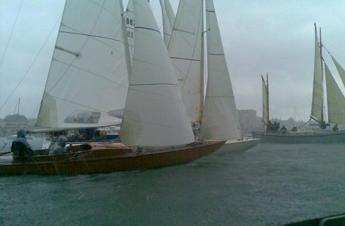 Several Classic Sailing Yachts with white sails compete in a race on a grey, rainy day with choppy water. People are visible on the boats, and buildings are faintly seen in the background through the mist.