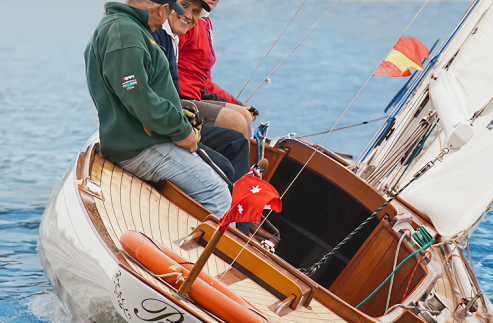 Three people are sitting and smiling on the deck of a Square Metre Yacht gliding on the water, with a red flag at the stern and a shoreline with trees and buildings blurred in the background.