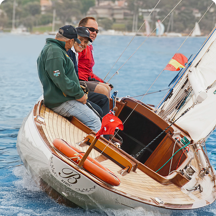 Three people are sitting and smiling on the deck of a Square Metre Yacht gliding on the water, with a red flag at the stern and a shoreline with trees and buildings blurred in the background.