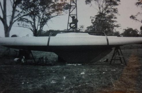 A black and white photo of a classic sailing yacht on land supported by stands, with a person sitting on top near a metal frame, surrounded by trees and grassy ground.