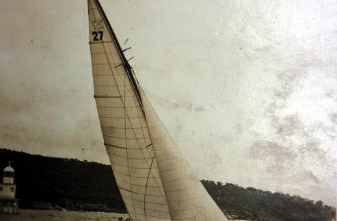 A vintage photo of a Classic Sailing Yacht labelled 27 gliding on the water with two people aboard. Hills and a small building are visible in the background. The word Aeolus is written on the photo’s upper edge.
