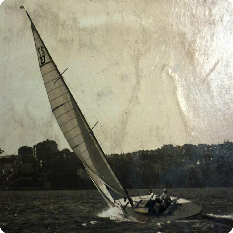 A vintage photo of a Schärenkreuzer sailboat with the number TS 27 on its tall sail, tilted sharply on a windy day, with several people aboard and a city skyline in the background.
