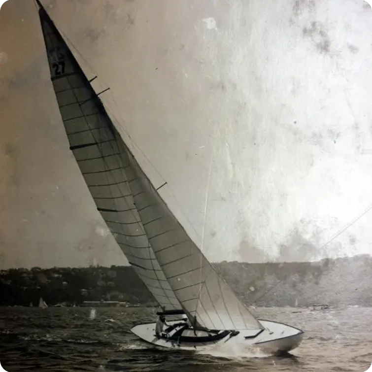 A vintage black and white photo of a Skerry Cruiser with a tall, angled mast gliding on the water, tilted by the wind. Hills and trees are visible in the background under a cloudy sky.