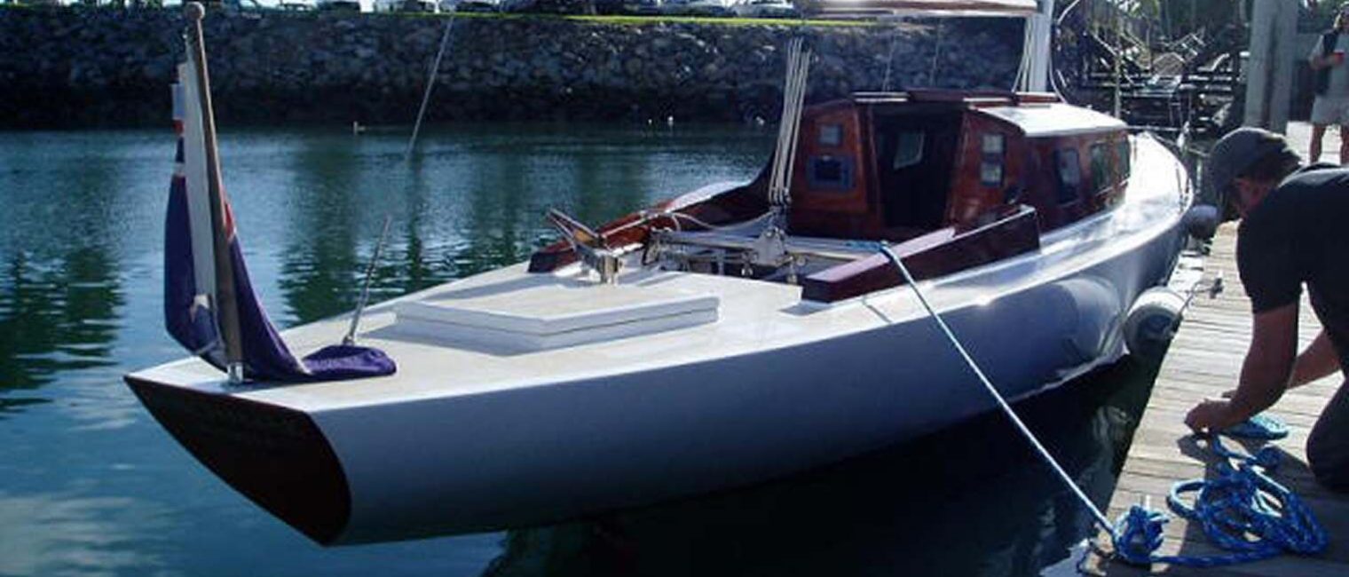 A sleek white sailboat, reminiscent of Classic Sailing Yachts, with a wooden cabin is moored by a pier. A person ties blue ropes to secure the boat. The calm water and rocky embankment frame parked cars in the background.