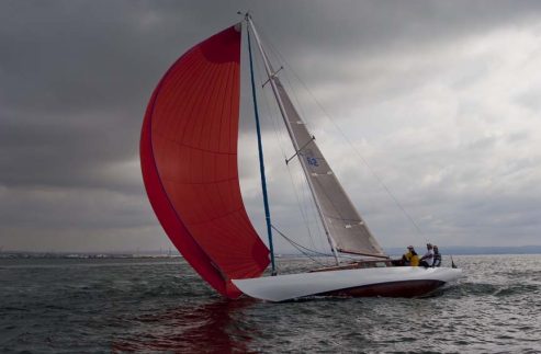 A sailing boat, reminiscent of Classic Sailing Yachts, with a large red spinnaker sail glides across the water under a cloudy sky. Several people are visible on board, and the coastline can be seen in the distance.