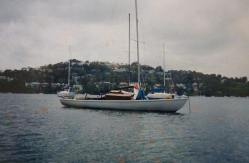 A white Schärenkreuzer is anchored on calm water with a hilly, tree-lined shoreline and houses in the background under a cloudy sky.