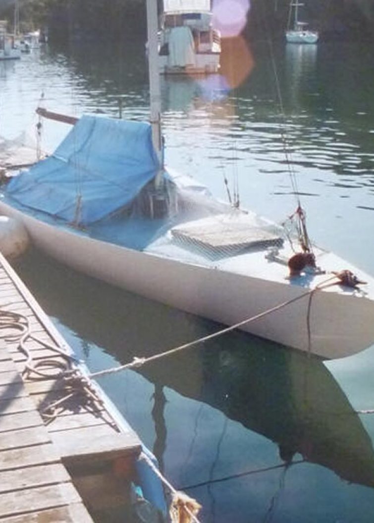 A small white Classic Sailing Yacht covered with a blue tarpaulin is tied to a wooden jetty in a calm marina, with several other boats and trees visible in the background.