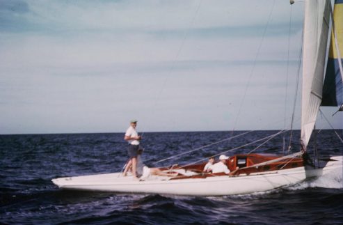 Three people are sailing on a sleek white Skerry Cruiser under a clear blue sky. One stands near the bow, while two relax beneath the cabin’s wooden structure. The calm water and classic Schärenkreuzer lines complete this serene scene.
