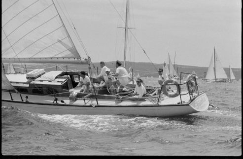 A group of people in casual clothing sail a classic sailing yacht on the sea, working together with the sails and ropes. Other Square Metre Yachts are visible in the background under a partly cloudy sky.