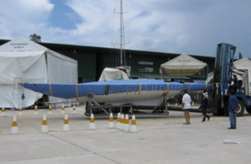 A long, narrow blue Skerry Cruiser is being transported on a trailer, with several people standing nearby. A forklift is positioned at one end, and tents and buildings are visible in the background.