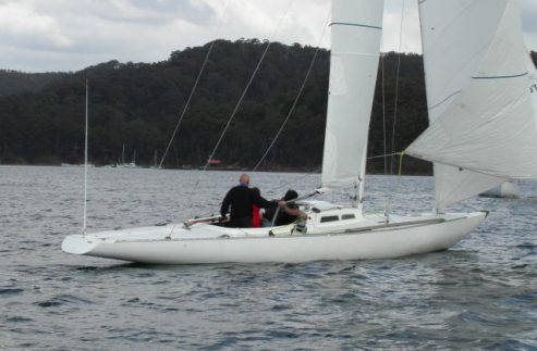 Two people are sailing a white Skerry Cruiser on a calm body of water, with forested hills in the background and cloudy skies overhead.