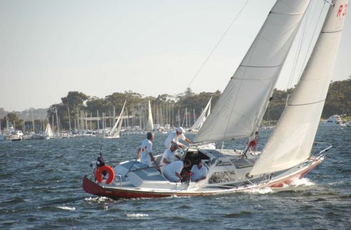 A small group of people sail a white and red Schärenkreuzer on a body of water, with many other sailing boats and trees visible in the background under a clear sky.