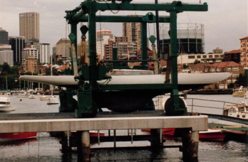 A classic sailing yacht is suspended on a green boat lift above a dock, with city buildings and moored boats visible in the background beside the waterfront.