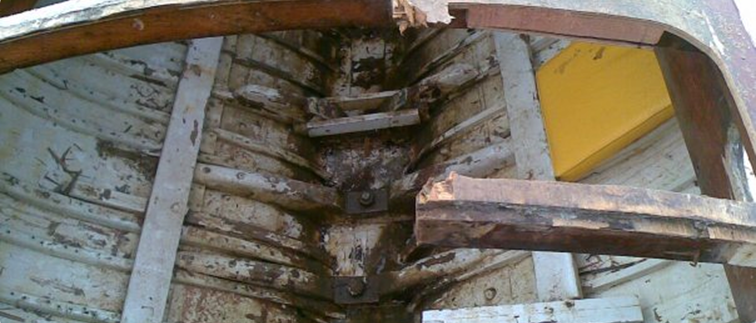 View of the damaged inside keel and ribs of an old wooden Square Metre Yacht, showing cracks and missing pieces in the hull structure, with worn paint and rust visible on the wood and metal parts.