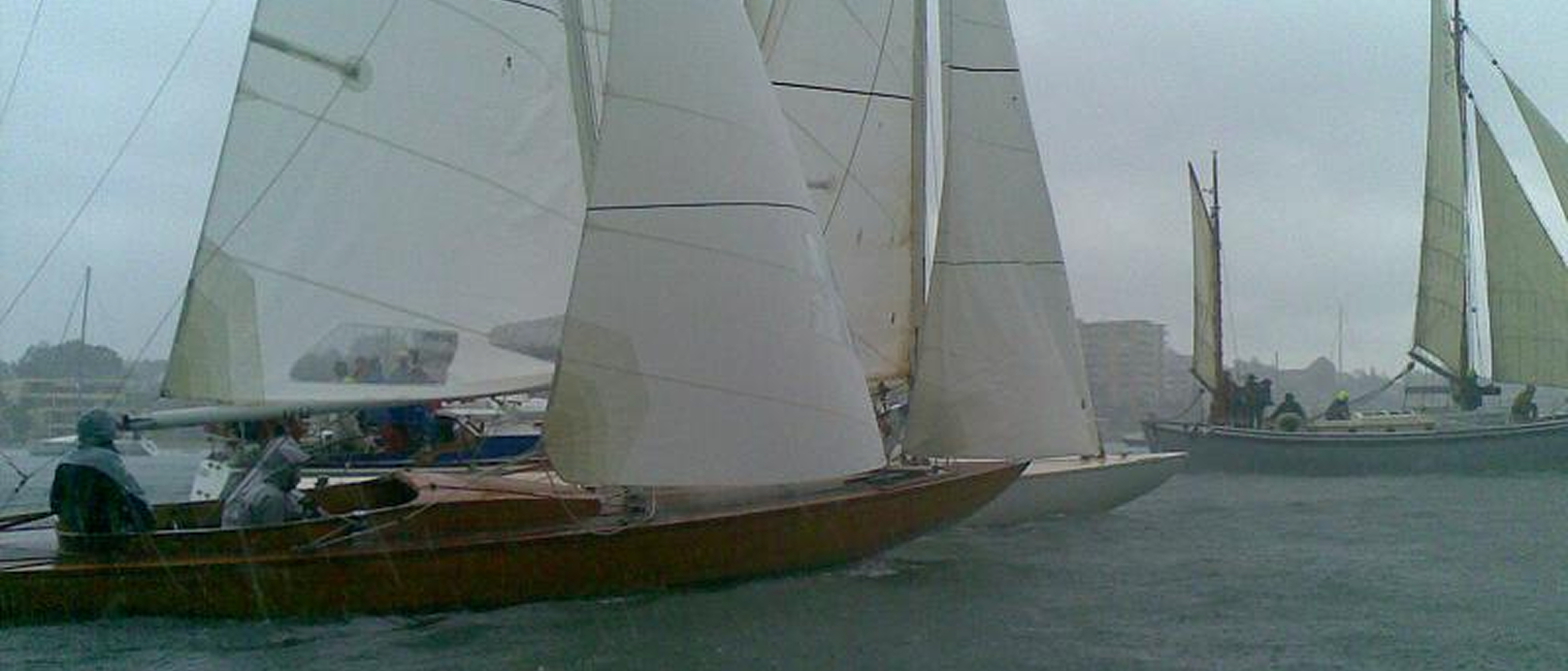 Several Classic Sailing Yachts with white sails race on a choppy, grey body of water in rainy, overcast weather. The scene appears misty, and people onboard are wearing rain gear. Buildings and trees are faintly visible in the background.