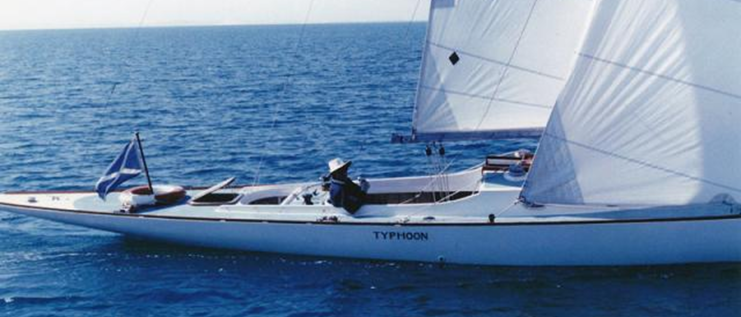 A sleek white Classic Sailing Yacht named Typhoon glides on calm blue water under clear skies. A person wearing a hat is seated at the helm, whilst a blue and white flag waves at the stern.