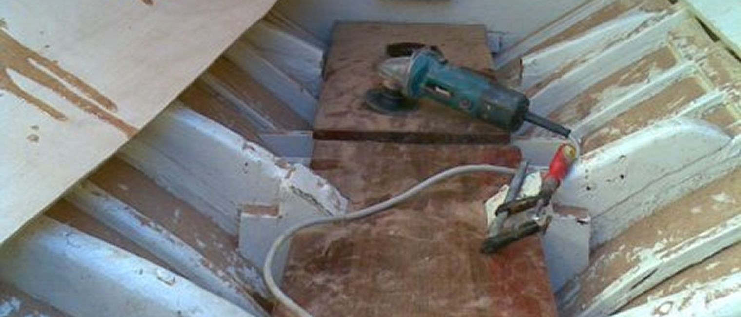 A partially repaired wooden boat interior with white framing, a power sander, and a clamp resting on wooden boards. Some tools and a partially detached plywood sheet are also visible—classic details reminiscent of Skerry Cruisers.