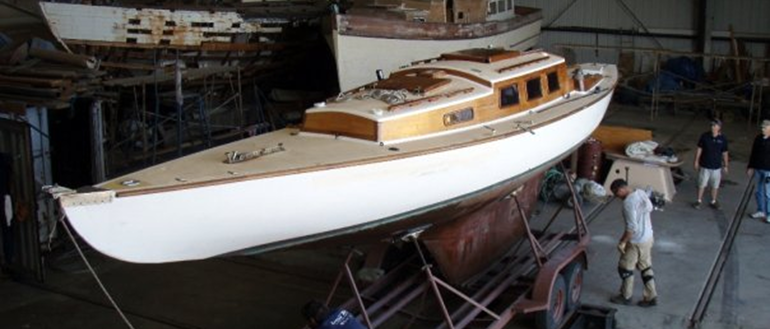 A classic Square Metre Yacht with a white hull and wooden cabin is being worked on in a large indoor workshop. Several people are around the boat, which is supported on a trailer. Other boats and tools are visible in the background.