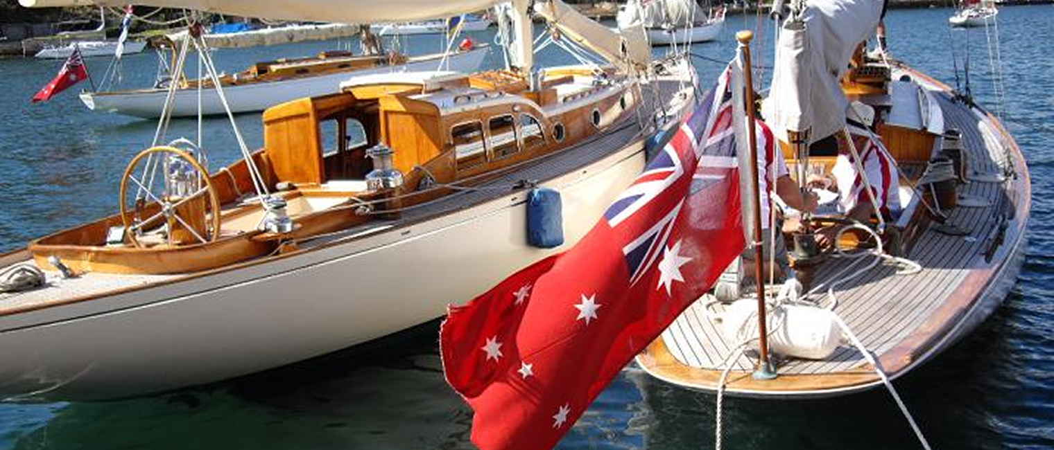 Two elegant Classic Sailing Yachts docked side by side on calm blue water, each displaying the Australian Red Ensign flag, with other boats and waterfront homes visible in the background.