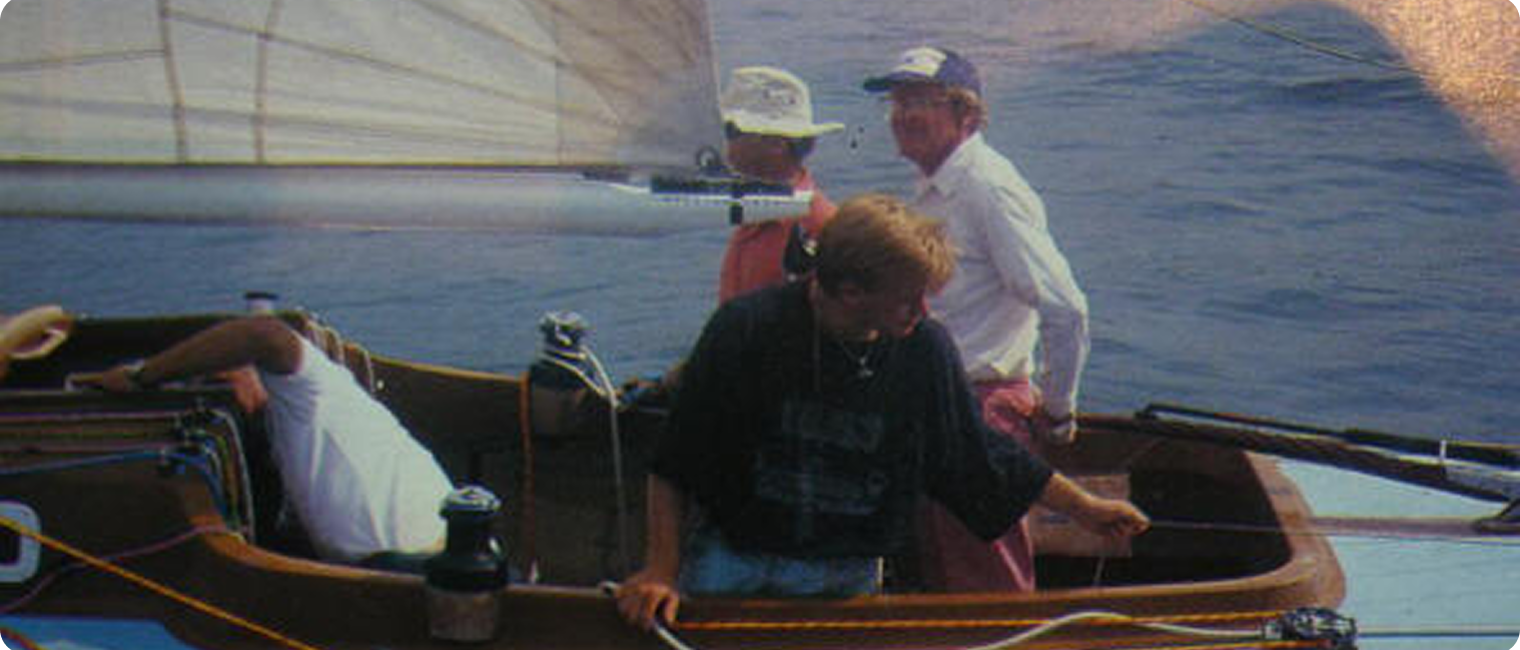 Four people are on a Classic Sailing Yacht at sea. One is seated and leaning over, whilst the others stand, handling ropes and looking out. The sails are up and the calm water reflects this timeless Skerry Cruiser experience.