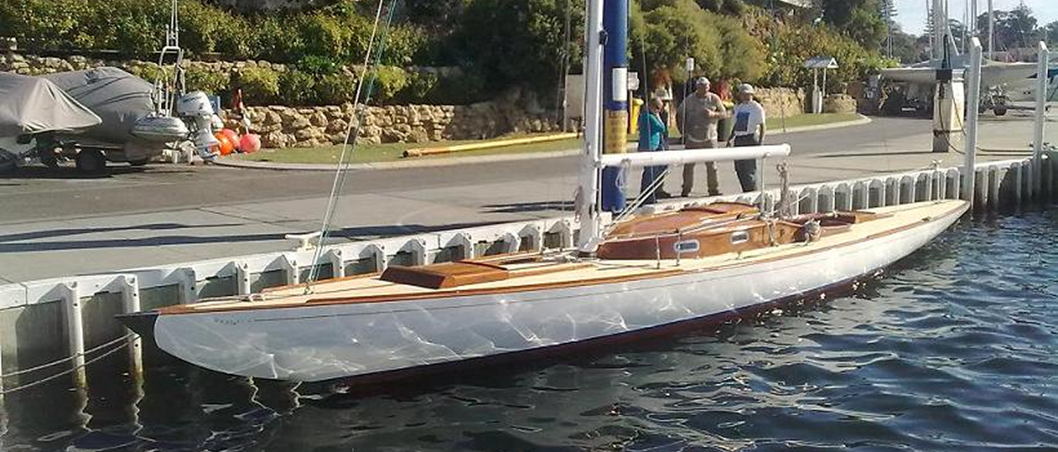 A sleek white wooden Schärenkreuzer sailboat with a polished deck is moored at a marina. Several people stand on the quay nearby, with greenery, stone walls, and classic sailing yachts in the background.