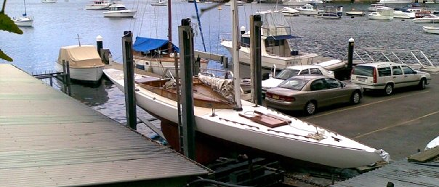 A white sailboat, reminiscent of Classic Sailing Yachts, is lifted on a boat hoist beside a car park, near a marina filled with boats and yachts on the water.