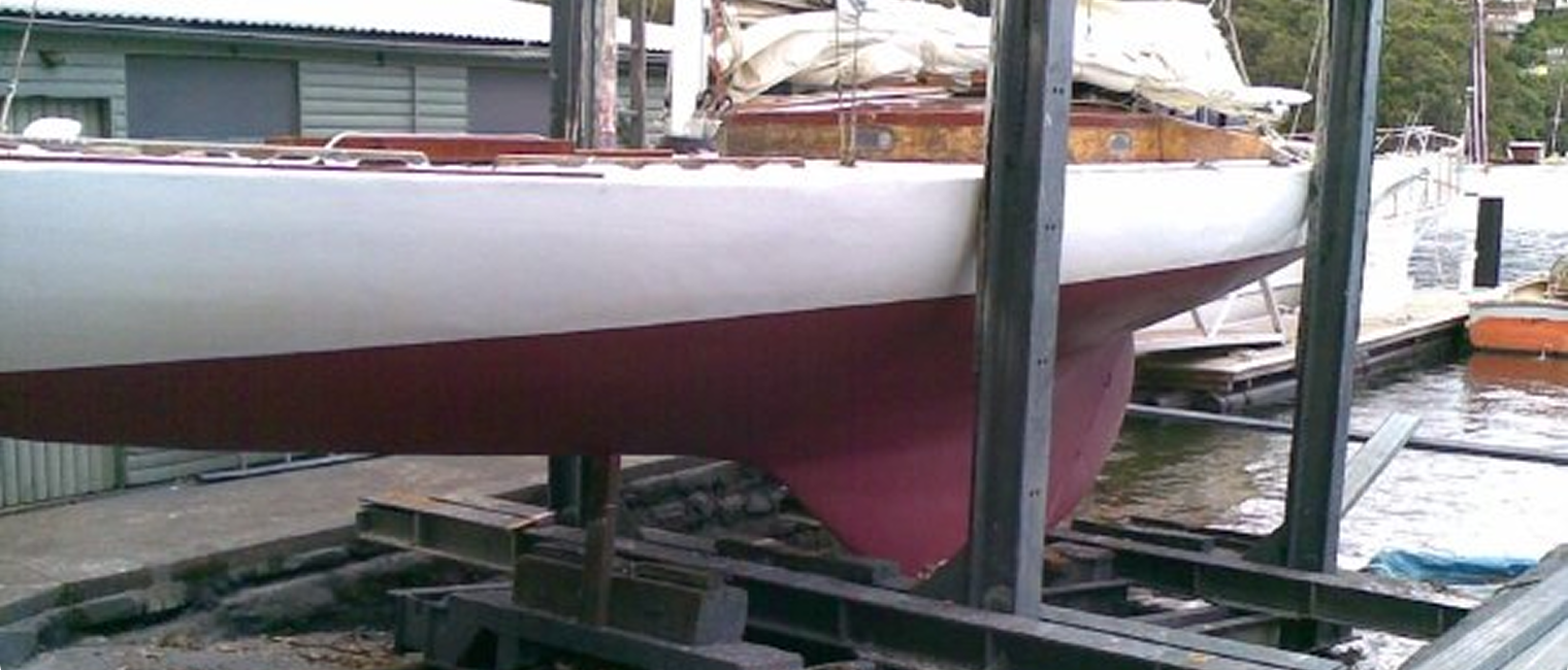 A white Classic Sailing Yacht with a red keel is lifted on a boat cradle at a quayside facility, with water and boats visible in the background.