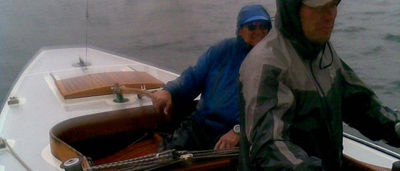 Two people in rain jackets are sailing a Skerry Cruiser on a cloudy day. One person is steering whilst the other sits behind, both appearing focused as they navigate the water.