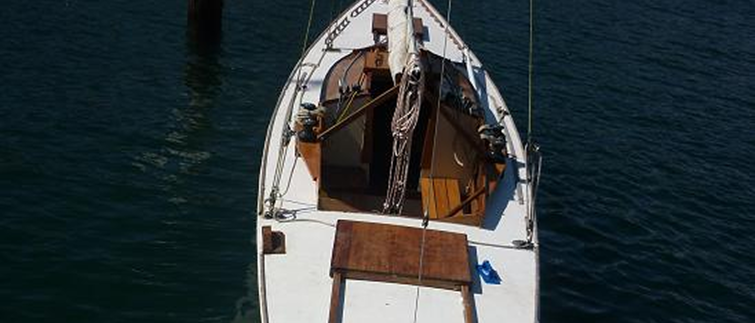 A white classic sailing yacht is moored on calm, dark blue water, viewed from above. The deck has wooden details, coiled ropes, and a table. The mast and rigging are visible, with a dock post nearby.