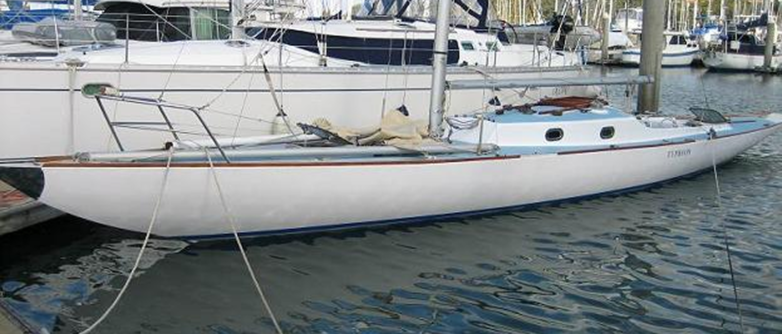 A white Schärenkreuzer sailboat is moored in a marina alongside other boats. It has a sleek hull, a covered cockpit, and is tied to the quay with ropes. The water is calm and classic sailing yacht masts are visible in the background.