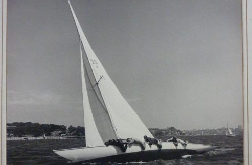 A vintage black-and-white photo of the sailboat Typhoon, a classic Square Metre Yacht, leaning dramatically on the water as crew members hang over the side to balance it. A distant shoreline and another sailboat are visible in the background.