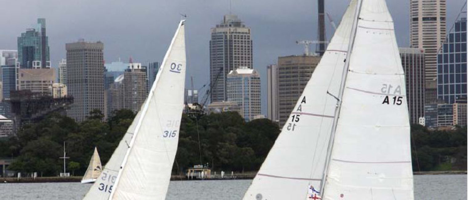 Two classic sailing yachts, known as Schärenkreuzer, race on the water with white sails. Several people are on board each vessel, while a city skyline with tall buildings and a tower rises in the background under cloudy skies.