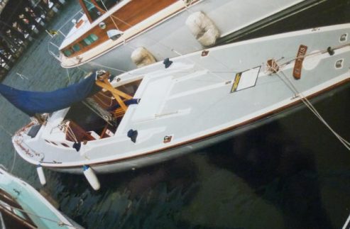 A white sailboat with a blue cover, reminiscent of classic sailing yachts, is moored at a marina alongside other boats. The deck is clean with minimal fittings, and fenders hang on the side to prevent damage. The water is calm and dark.