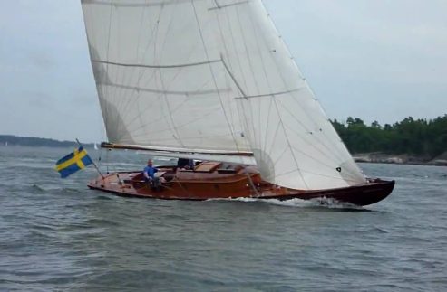 A classic wooden sailboat, reminiscent of Classic Sailing Yachts, with white sails and a Swedish flag glides across choppy waters. Two people are on board as the vessel passes a forested shoreline in the background.