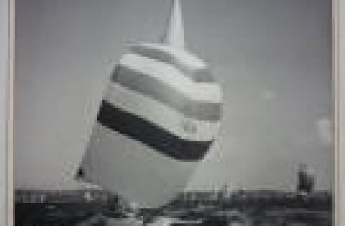 A classic sailing yacht with a large striped spinnaker sail glides across the water on a sunny day, with other Square Metre Yachts visible in the background.