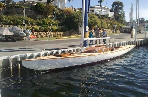 A sleek wooden Schärenkreuzer sailboat with a tall mast is moored at a marina beside the quay, with several people near the waterfront and houses and greenery in the background.