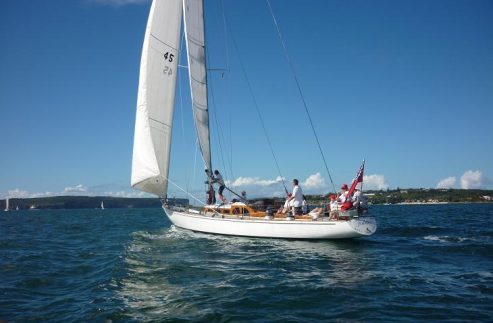 A classic Skerry Cruiser with several people on board sails on blue water under a clear sky. Land with green trees is visible in the background. The elegant yacht has raised white sails and a red flag at the stern.
