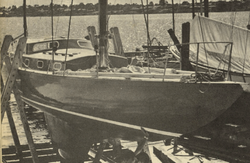 A Skerry Cruiser is moored on a wooden slipway near the shore, with rigging and masts visible. Calm water and a distant tree-lined shoreline are in the background under a bright sky.